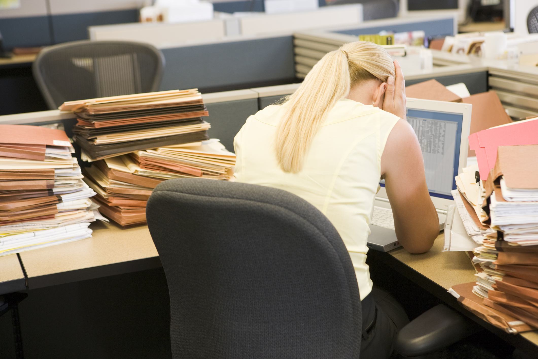 Businesswoman sitting with her head in her hands in cubicle with laptop and stacks of files