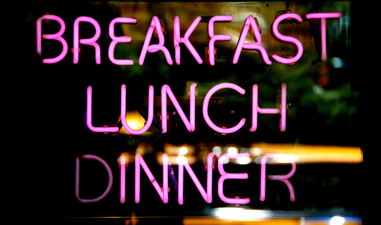 A pink neon sign in a diner window reading "Breakfast, Lunch, Dinner".