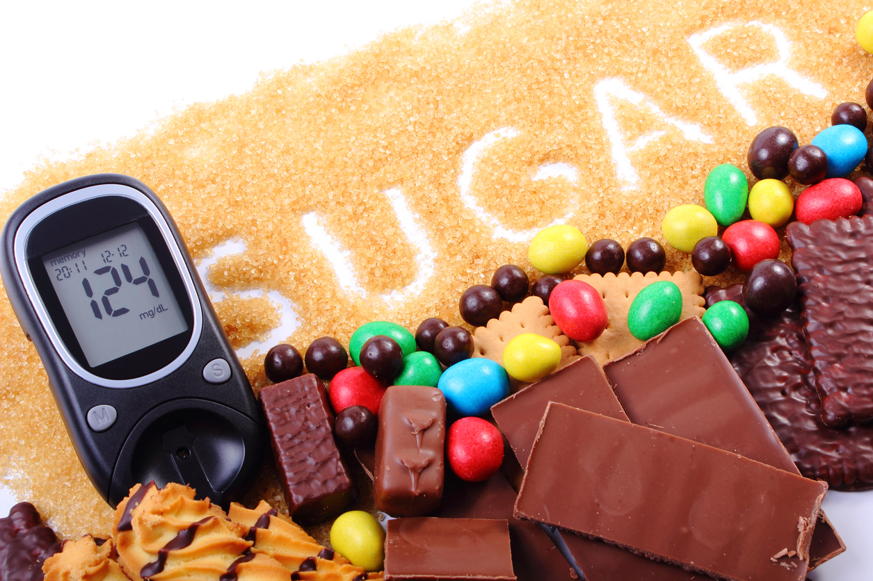 Glucose meter, granulated natural brown cane sugar and a lot of candies and cookies, concept of too many sweets, unhealthy food and diabetes