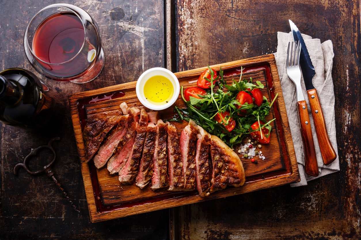 Sliced grilled beef barbecue Striploin steak and salad with tomatoes and arugula on cutting board and wine on dark background