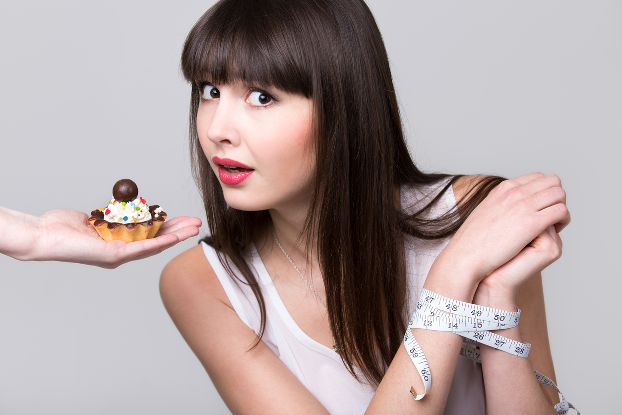 Young dieting woman with hands tied with measurement tape sitting in front of tart cake, got caught while trying to reach it and take a bite, studio, gray background, isolated