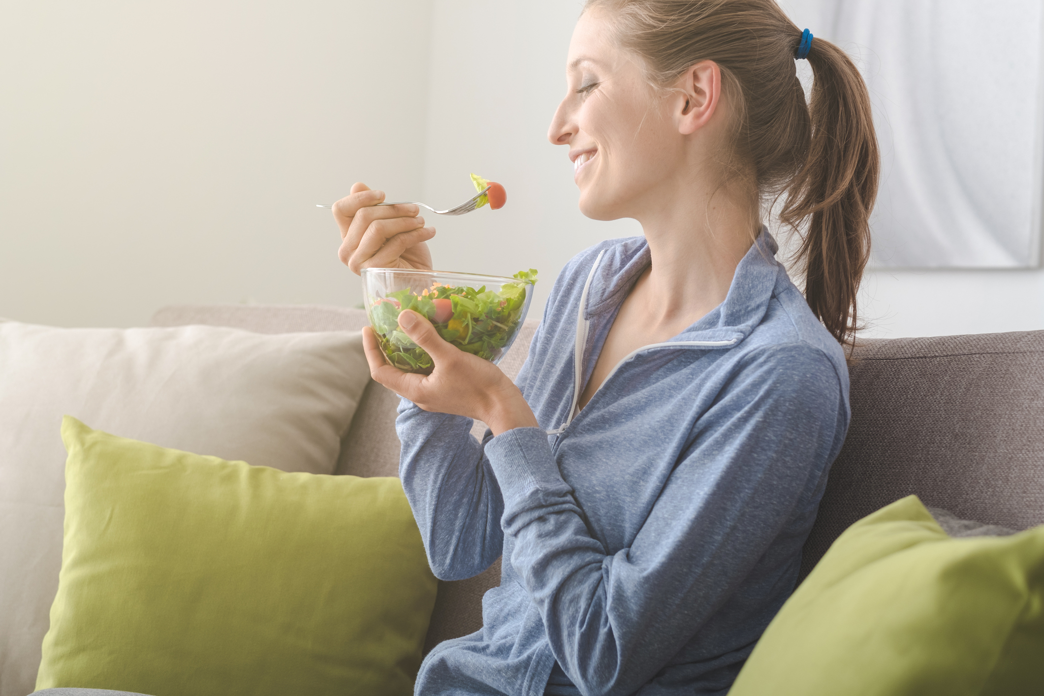 Young blonde woman relaxing on the couch at home and eating a fresh garden salad, healthy lifestyle and nutrition concept