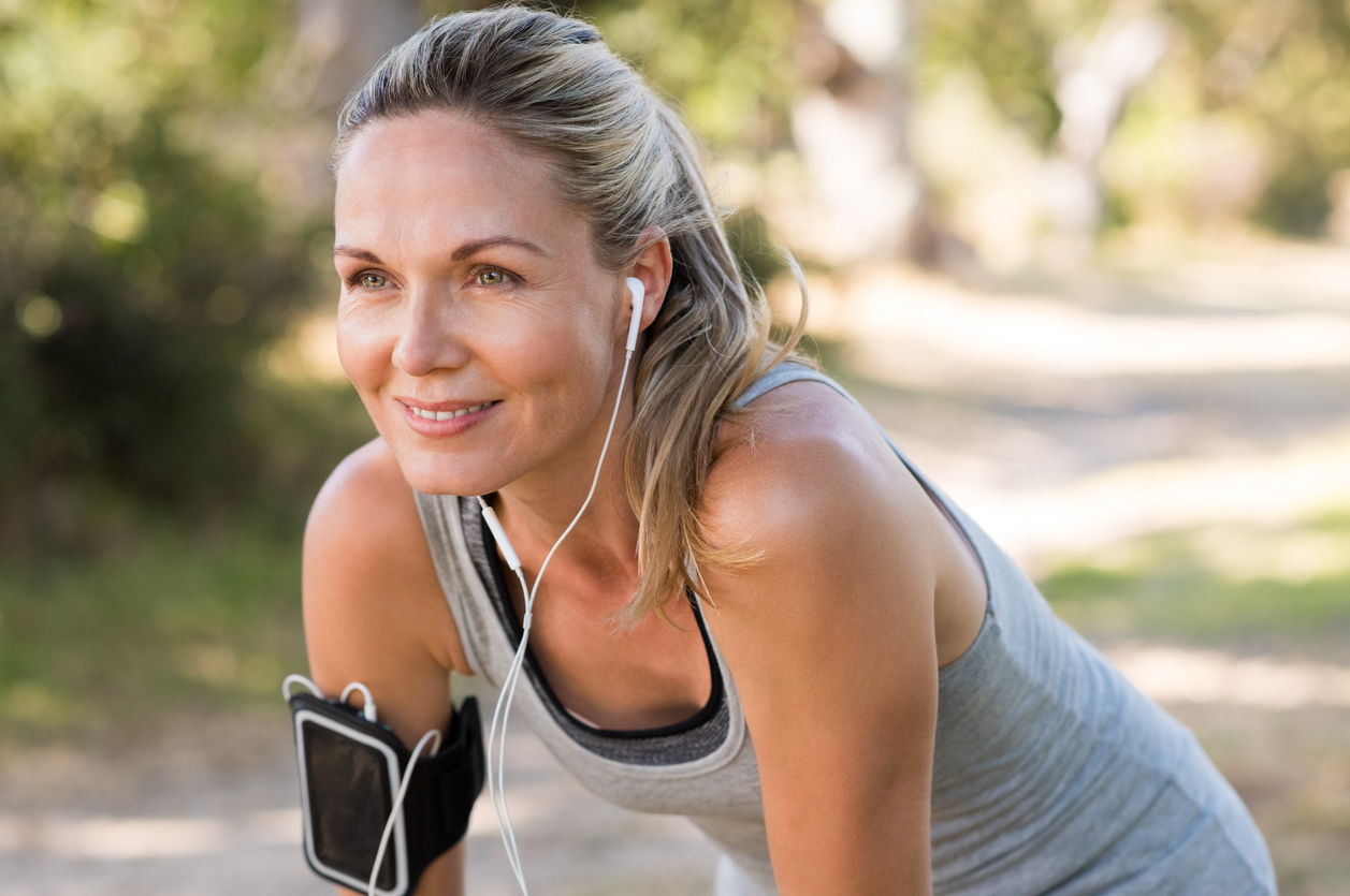 Portrait of athletic mature woman resting after jogging. Beautiful senior blonde woman running at the park on a sunny day. Female runner listening to music while jogging.