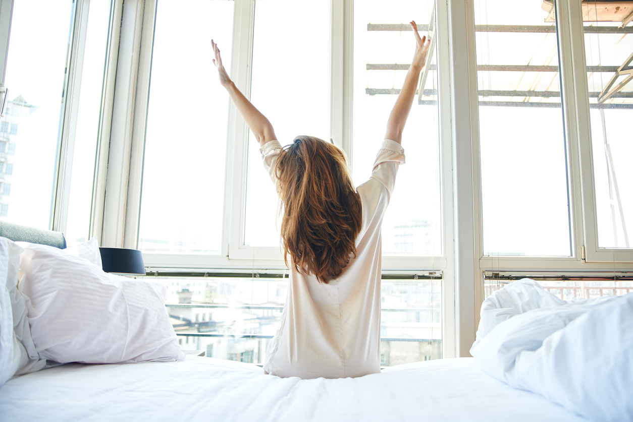 Young woman is doing morning stretching in bed, arms raised, rear view