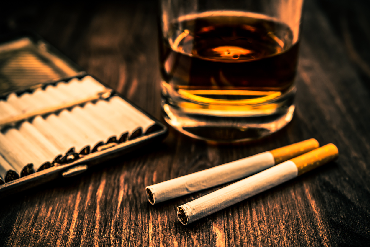 Glass of whiskey and two cigarettes with a cigarette case on a wooden table. Focus on the cigarettes, shallow depth of field, image vignetting and the orange-blue toning