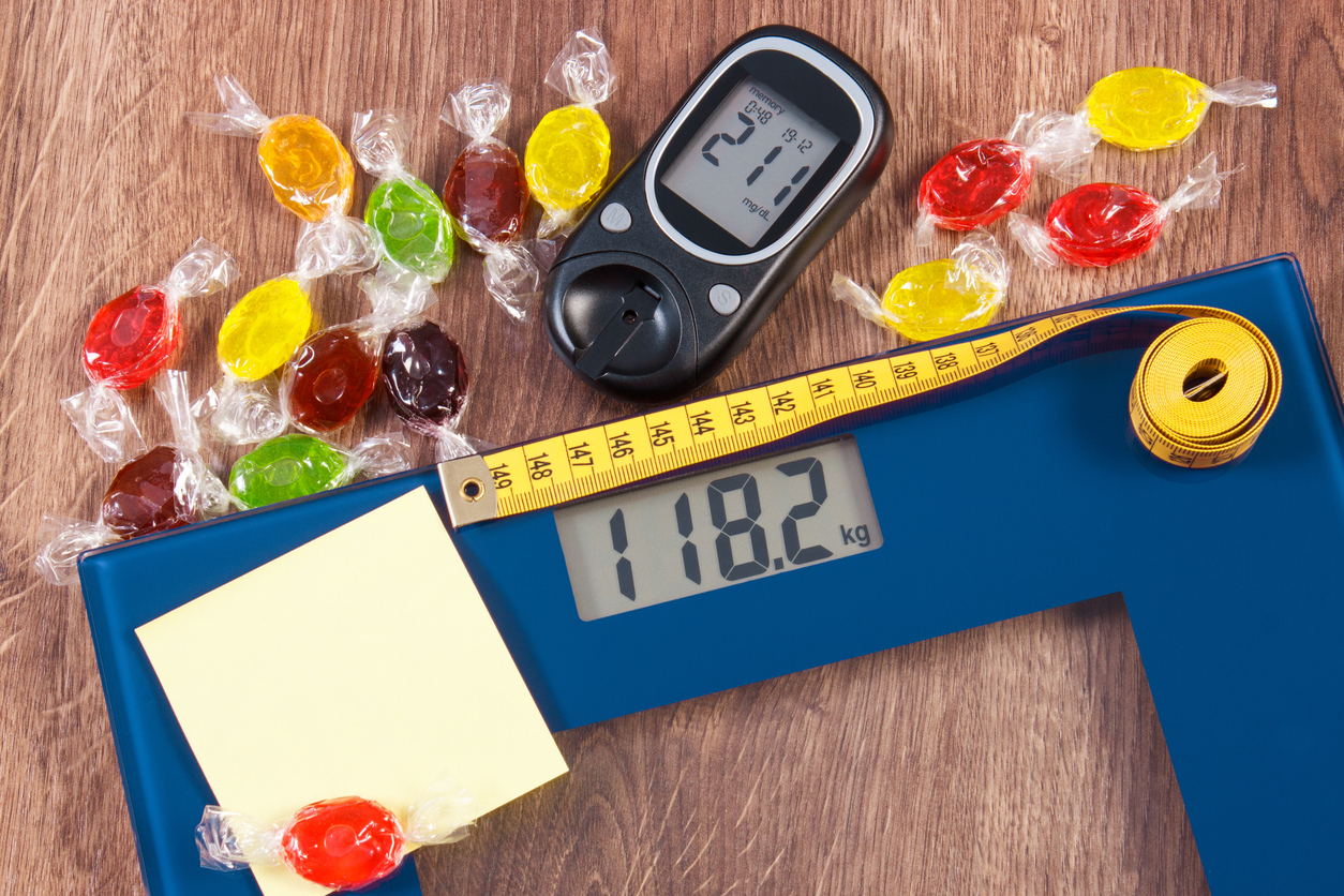 Electronic bathroom scale and glucose meter with high result of measurement weight and sugar level and heap of colorful candies, diabetes, slimming and reduction eating sweets, copy space for text
