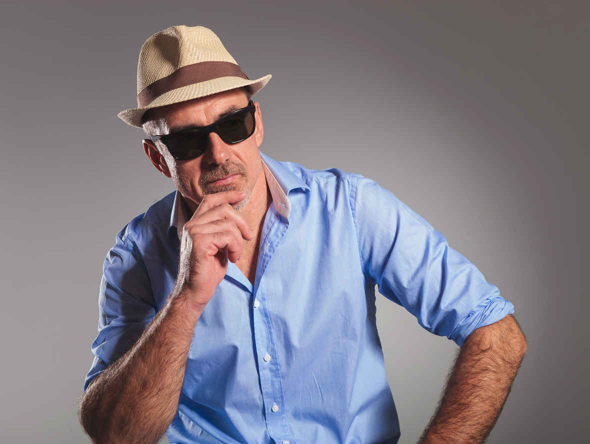 closeup portrait of mature man in open shirt thinking and looking away from the camera. the man is wearing brown hat and black sunglasses in gray studio background.