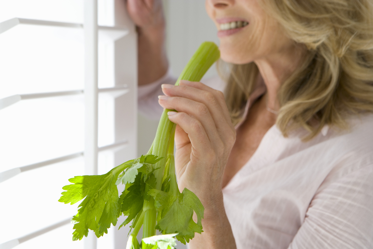 Mature woman holding celery stick, standing by window, smiling, side view