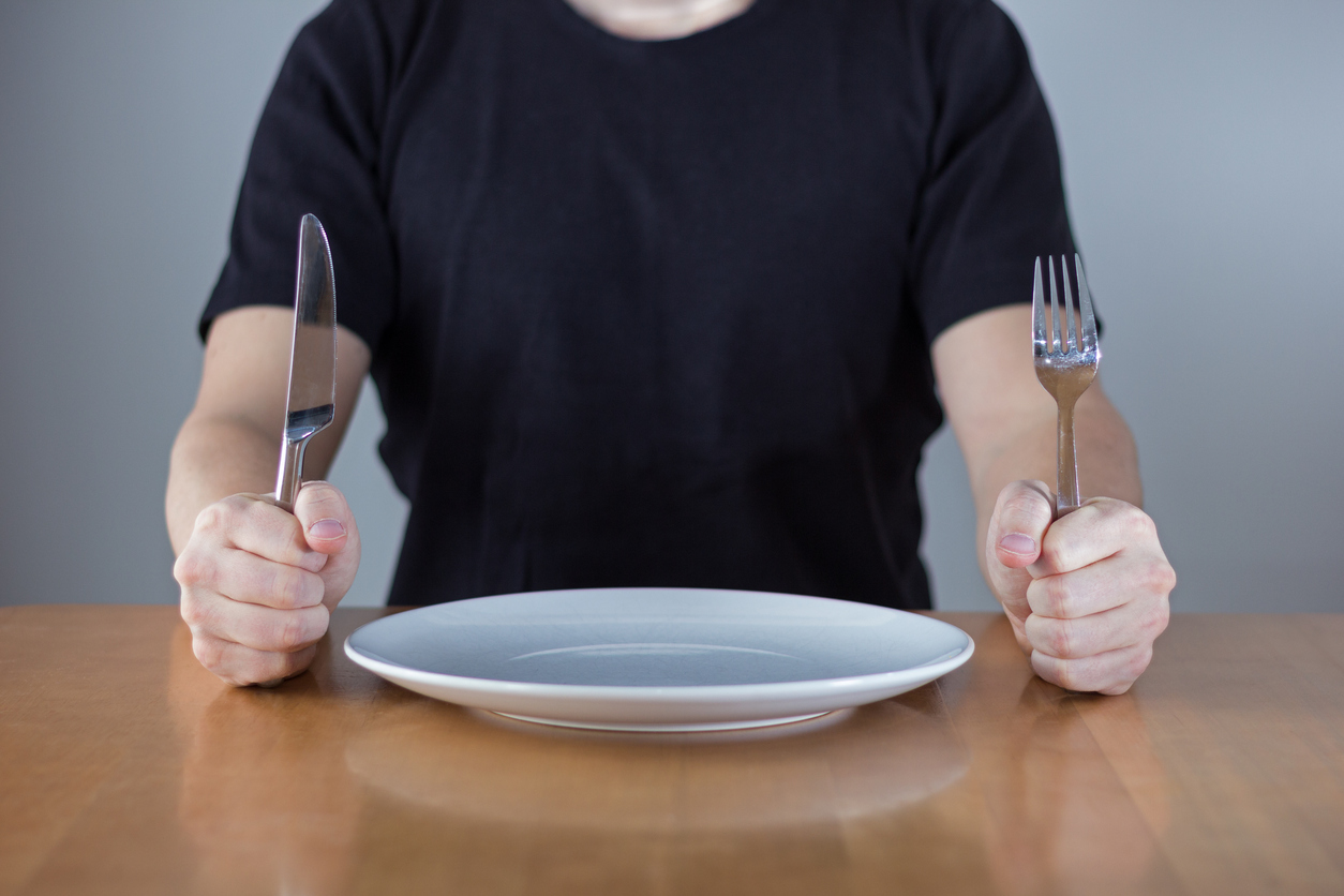 An unrecognizable man wearing black shirt sitting at a table in front of an empty plate waiting for food, holding fork and knife in his hands.