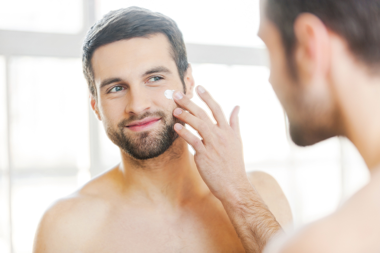 Handsome young shirtless man applying cream at his face and looking at himself with smile while standing in front of the mirror