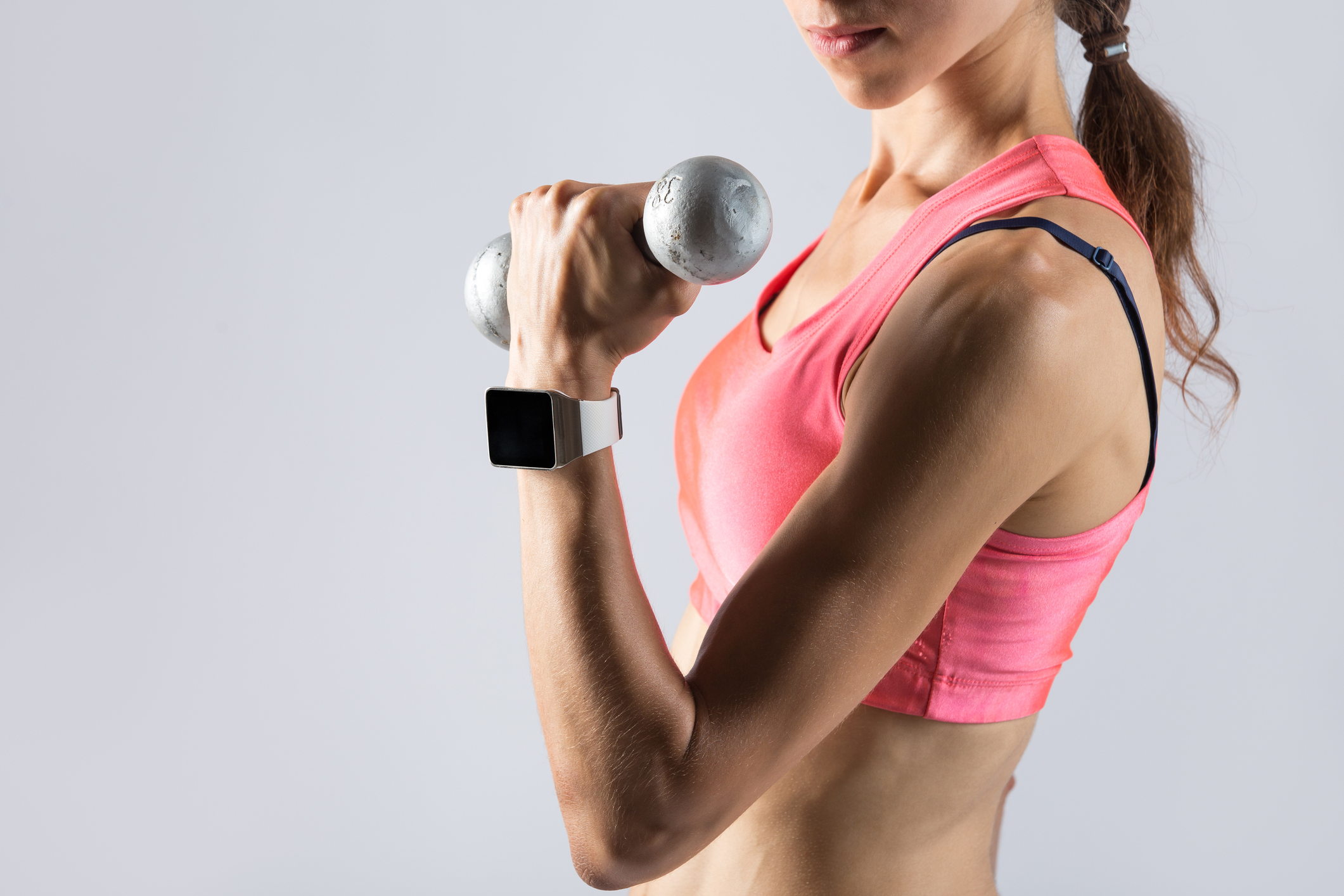Close-up of torso of beautiful young fitness person wearing smartwatch and red sportswear top holding dumbbell. Sporty model girl working out, doing weight training with dumbbells on grey background