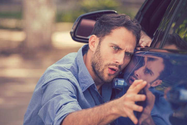 Frustrated upset young man looking at scratches and dents on his car outdoors